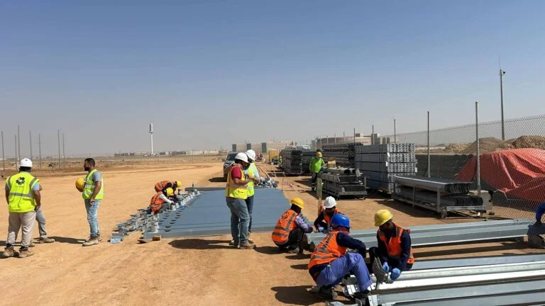 Workers assembling shade house steel framing at a sandy Saudi construction site